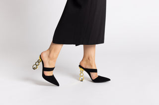 Black suede pointed closed toe Kat Maconie mule, with a-symmetrical strap over arch of foot stacked on 3 crystal gold heels with women's legs and black skirt