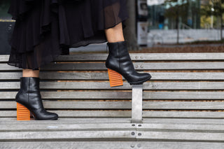 United Nude, Hi Rise, Black leather patent boot with zip and black sole thread with a see through orange heel styled with black dress on a bench seat and modelled with feet and legs , The Shoe Curator