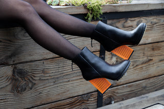 United Nude, Hi Rise, Black leather patent boot with zip and black sole thread with a see through orange heel styled with sheer leggings and modelled with feet and legs, The Shoe Curator