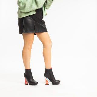 United Nude, Glam Bootie, Black patent ankle boot with pink and red lightning bolt heel with silver stability styled with black mini skirt and green jersey modelled with feet and legs, The Shoe Curator