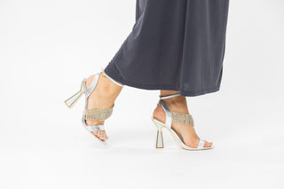 Kat Maconie, Mila, Silver leather buckle strap with crystal fringe down the foot with hourglass heel with gold edging styled with dark grey dress and modelled with legs and feet, The Shoe Curator