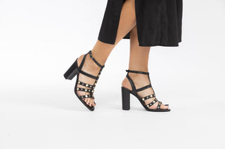 Ted Baker, Carolya, Black leather pump with gold dot detailing with straps up and down the ankle with block heel styled with black dress and modelled with feet and legs, The Shoe Curator