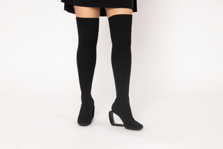 United Nude, Mobius Long Boot Hi, Black mid thigh boot with black stretchy material, heel with a thin piece creating a hollow middle styled with black mini dress and modelled with feet and legs, The Shoe Curator